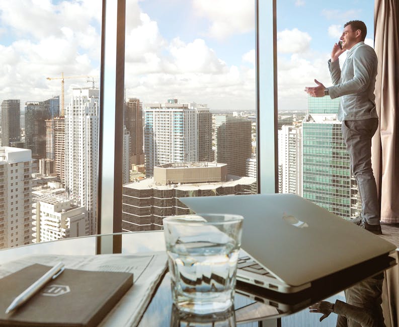 Businessman talking on smartphone by window with city skyline view, laptop and glass in foreground.